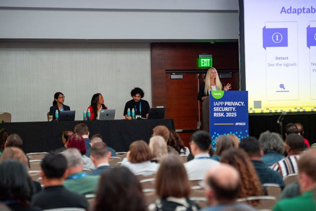 Conference session with a speaker standing at a podium labeled “IAPP Privacy. Security. Risk. 2025” in front of a large screen displaying a slide titled “Adaptability” with bullet points including “Detect: See the signals.” Three panelists are seated at a long table with laptops and water bottles. Audience members are seated in rows facing the stage.