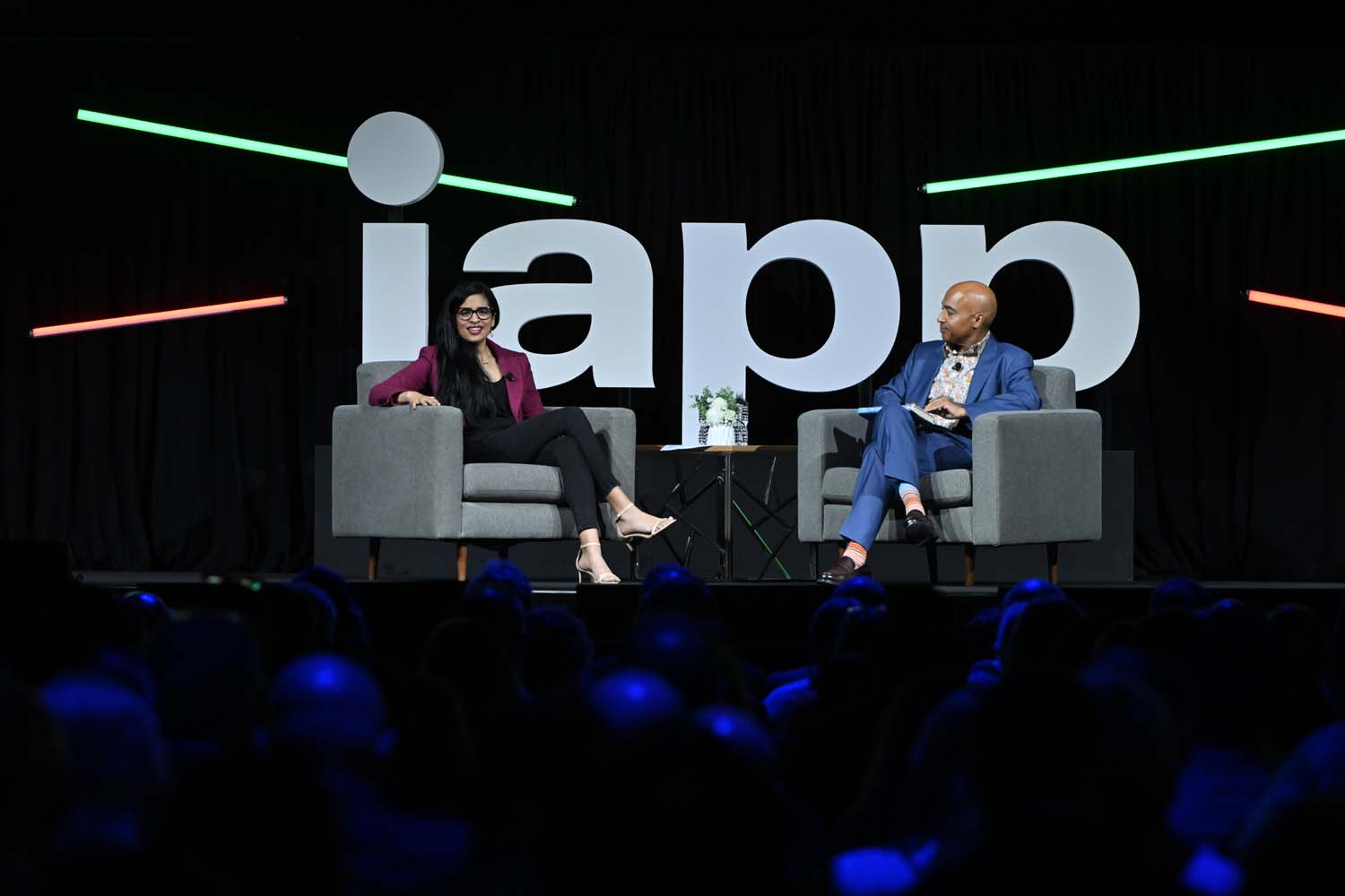 Two speakers sit in upholstered chairs on a stage beneath oversized IAPP lettering, facing each other across a small table during a live onstage discussion.