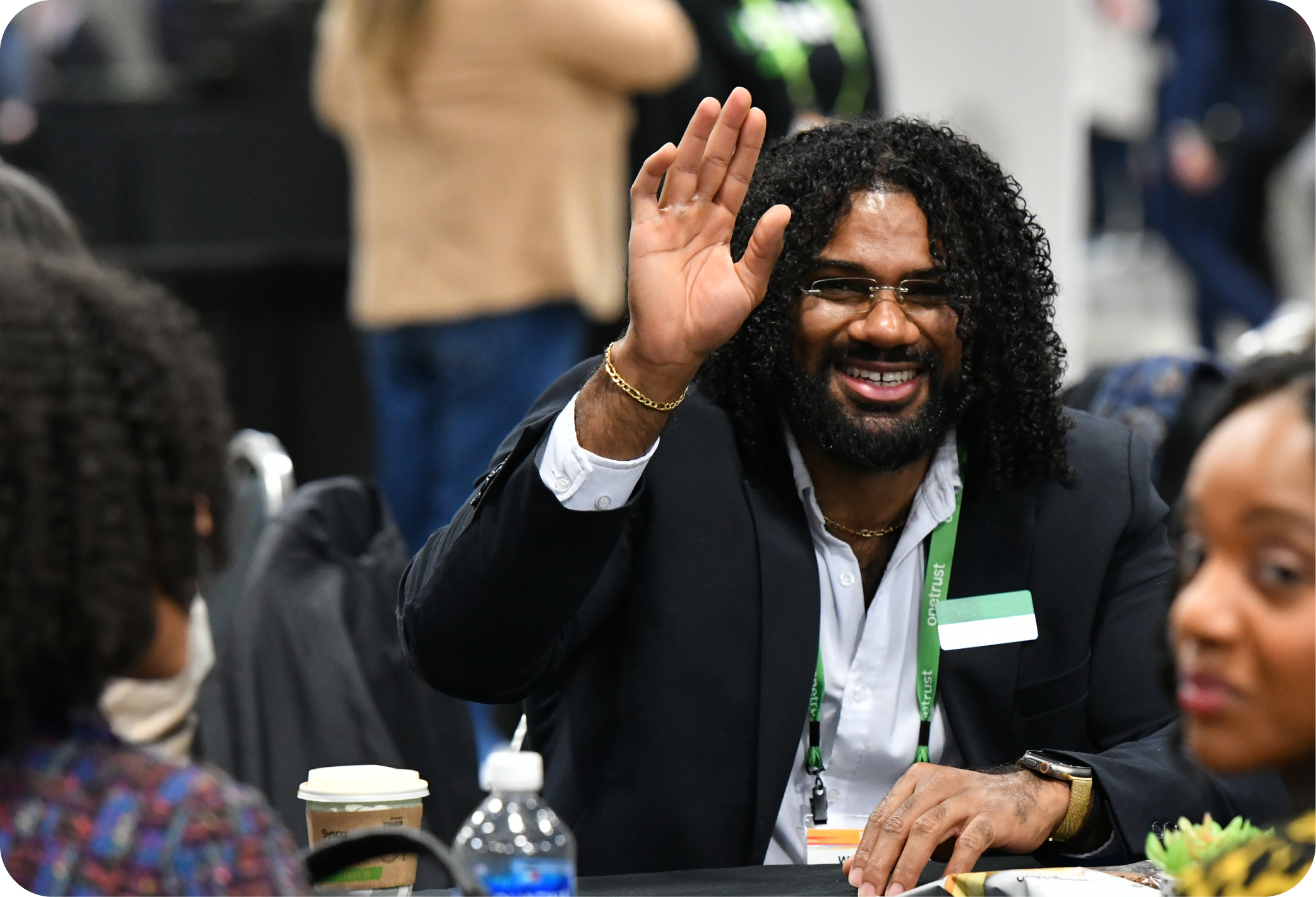 an african american man in a suit jacket is sitting a table and raising his hand and smiling