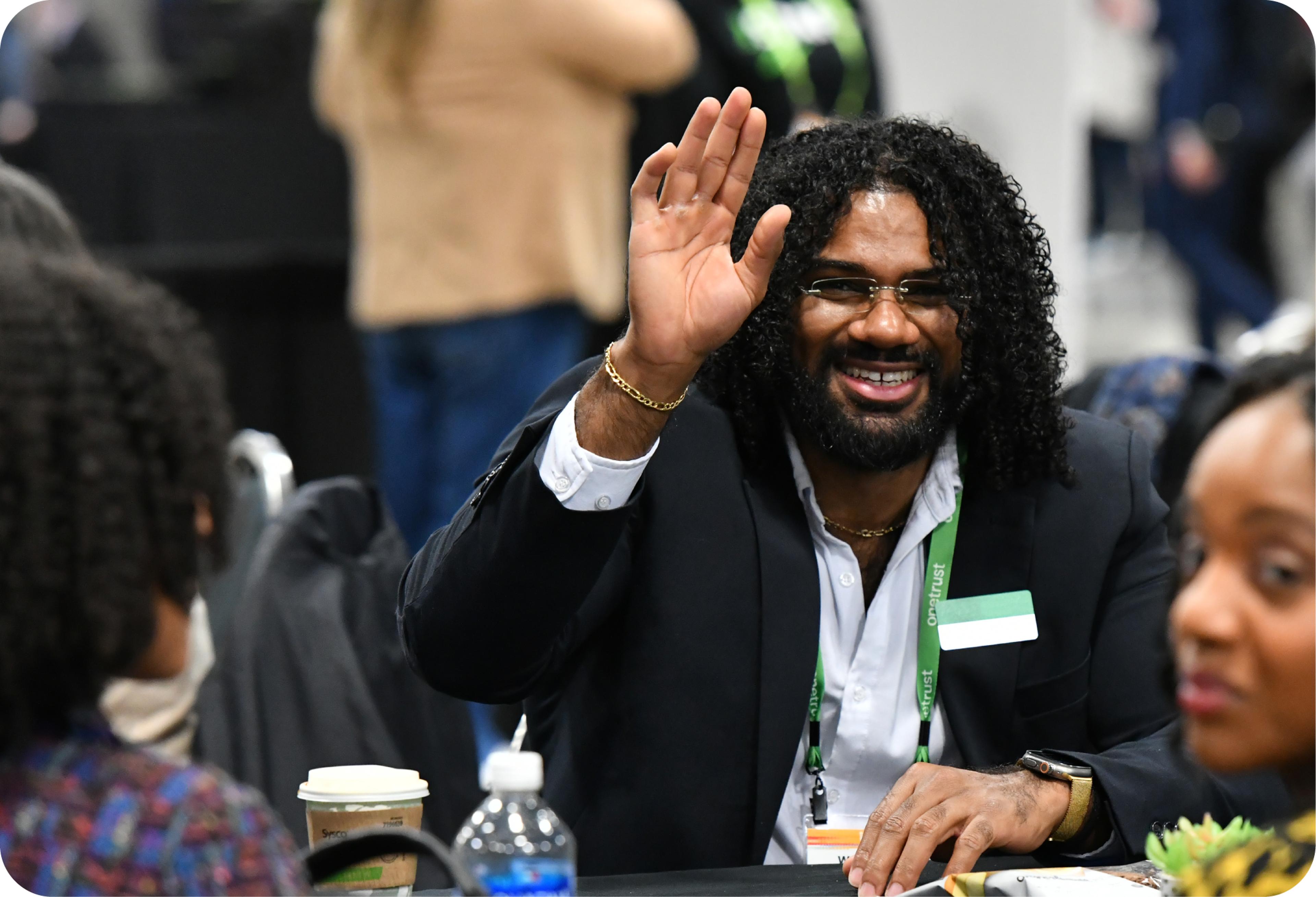 an african american man in a suit jacket is sitting a table and raising his hand and smiling