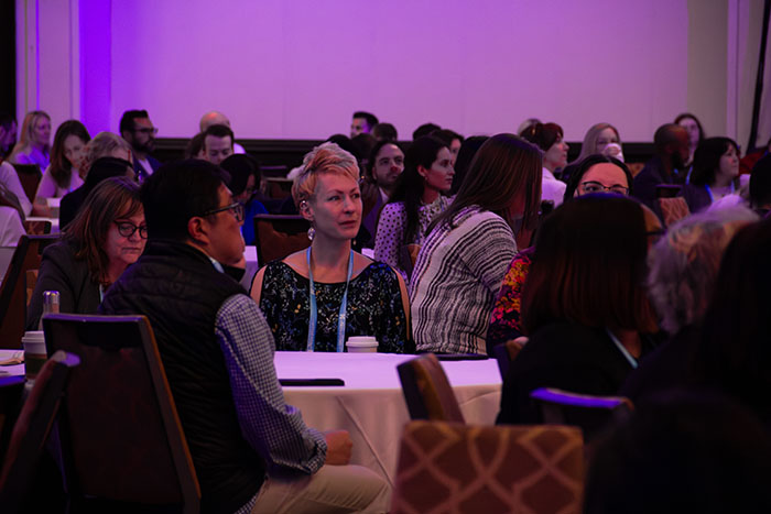 Audience seated at round tables in a conference room, listening to a presentation with purple lighting and multiple attendees facing toward the unseen stage.