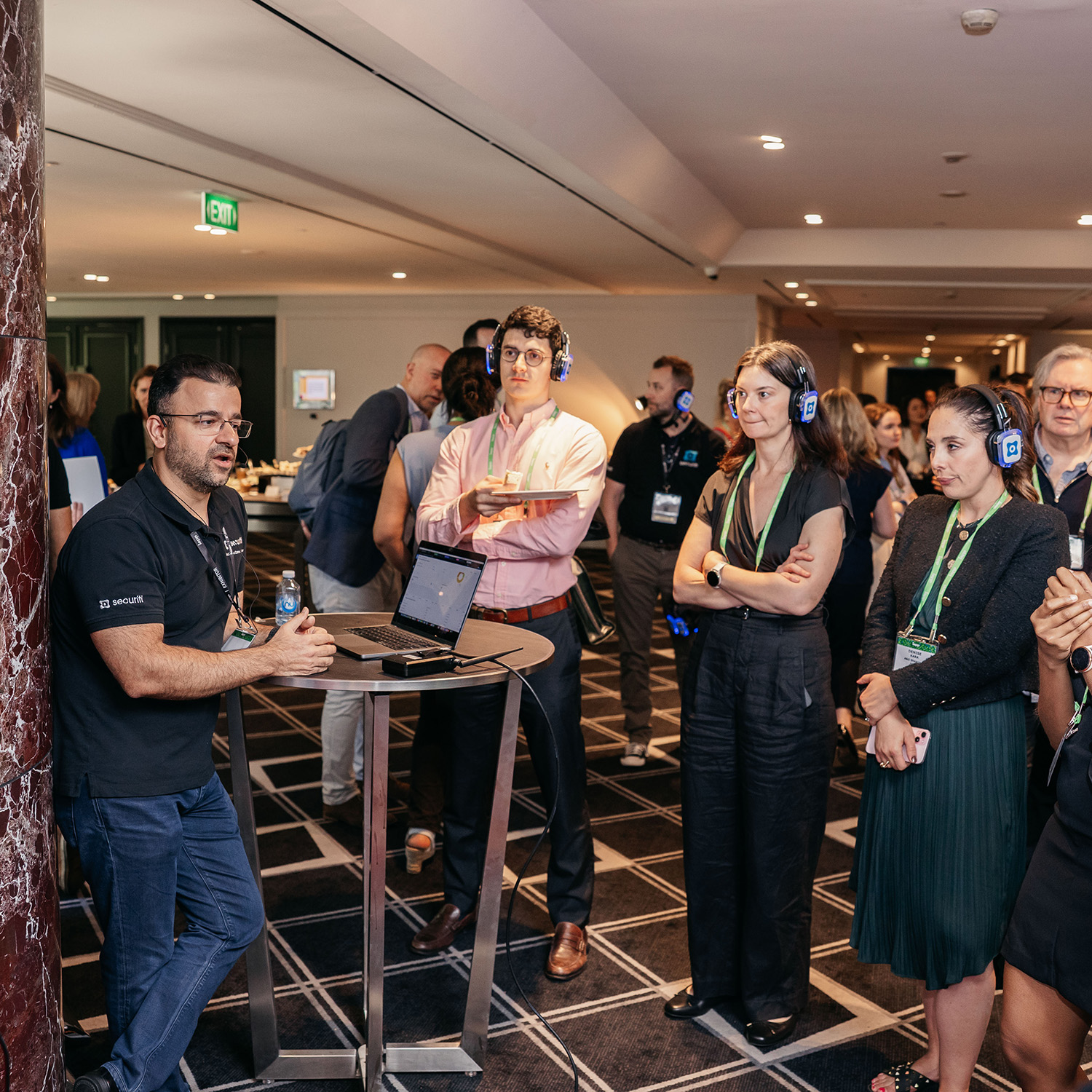Group gathered around a small table in a networking area, listening to a presenter with a laptop and screen.