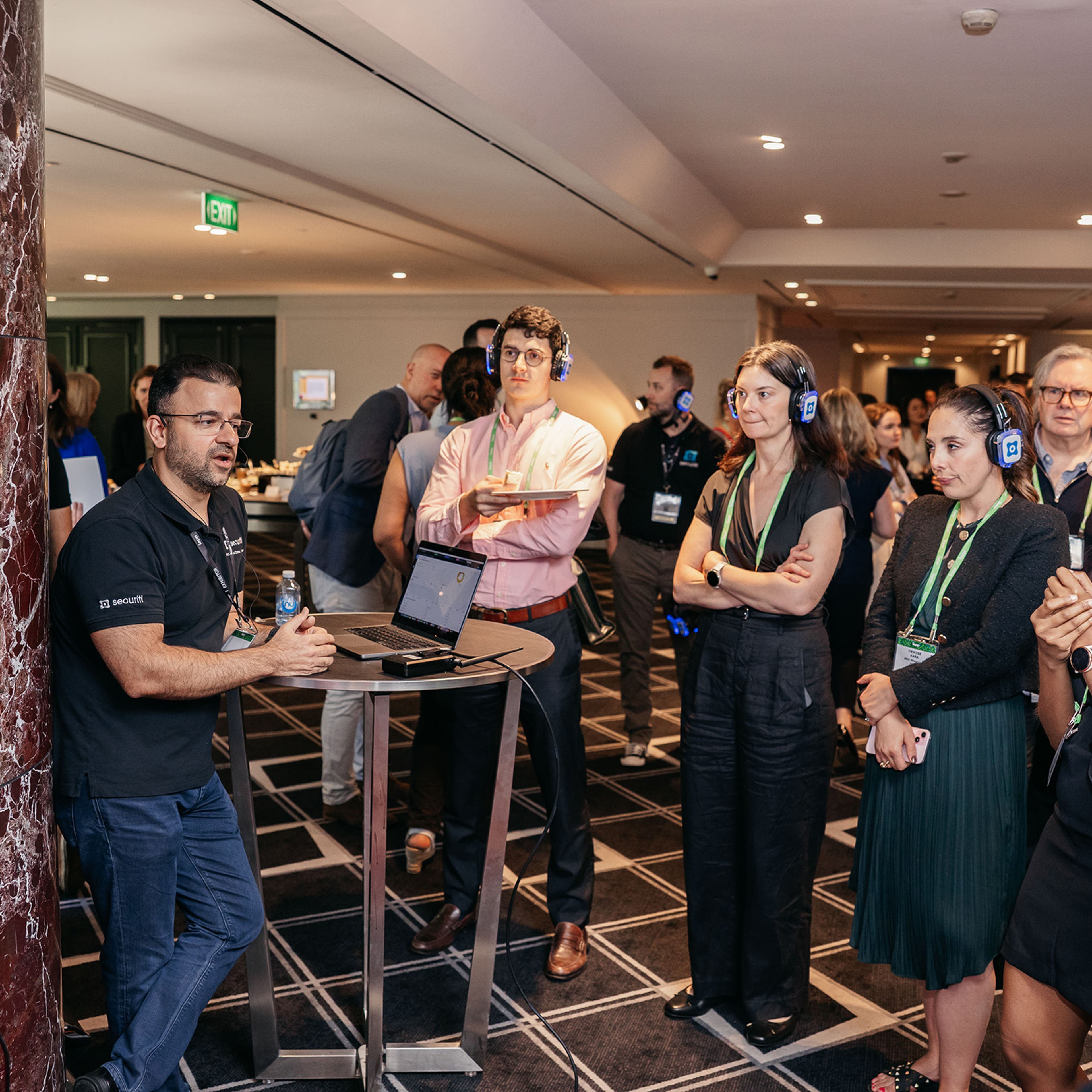 Group gathered around a small table in a networking area, listening to a presenter with a laptop and screen.