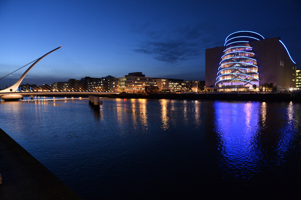 Nighttime view of the Samuel Beckett Bridge and the Convention Centre Dublin along the River Liffey. The bridge features a harp-like design with white cables, and the convention center is illuminated with bright blue curved light bands. Reflections of the lights shimmer on the water under a deep blue sky.