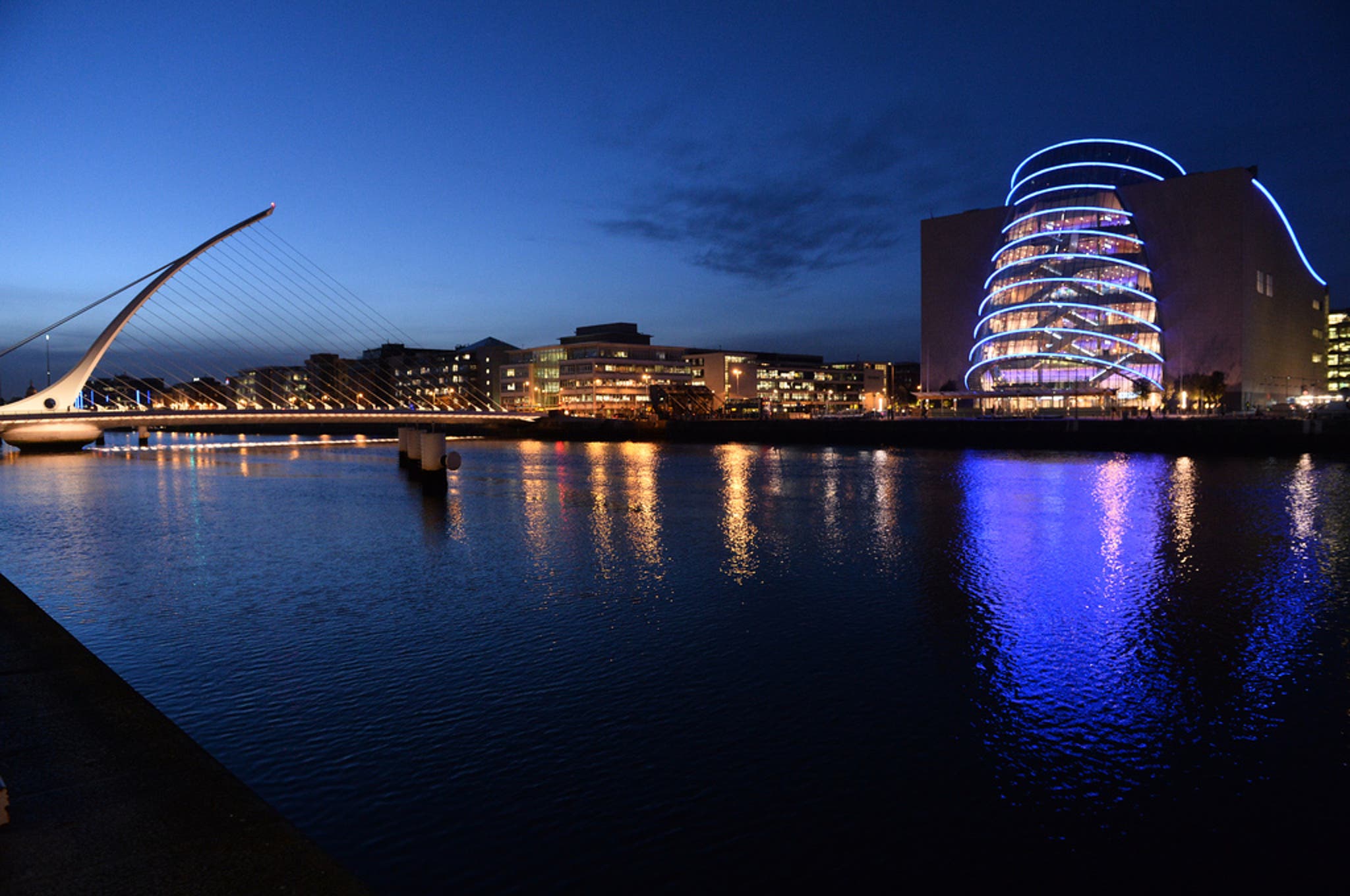 Nighttime view of the Samuel Beckett Bridge and the Convention Centre Dublin along the River Liffey. The bridge features a harp-like design with white cables, and the convention center is illuminated with bright blue curved light bands. Reflections of the lights shimmer on the water under a deep blue sky.