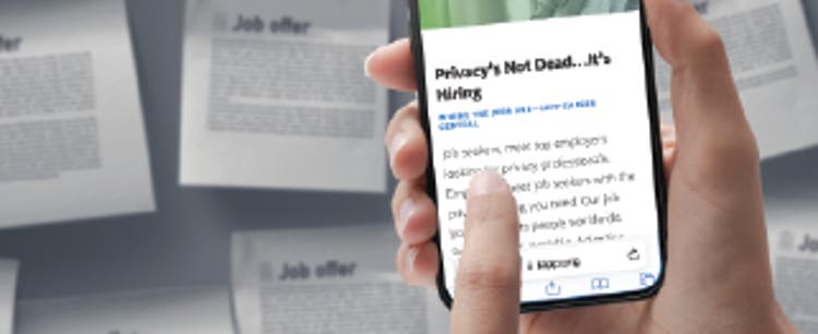 A closeup of a person holding a mobile phone with the screen displaying the IAPP Career Central page, which reads, "Privacy's Not Dead... it's Hiring." A job board is out-of-focus in the background, with several papers tacked to the board that read "Job offer" in big letters and paragraphs of text below it.