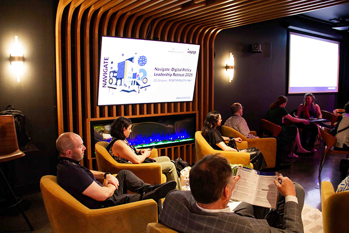 Small group seated in a lounge-style meeting area with curved wooden paneling and two large screens displaying “Navigate: Digital Policy Leadership Retreat 2025.”