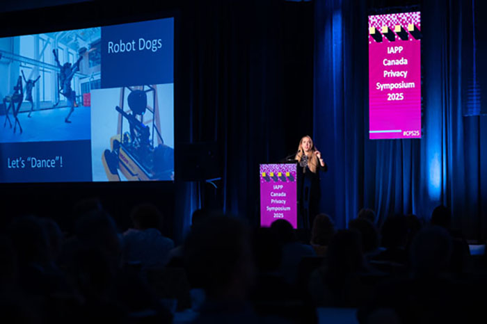 Presenter on stage speaking to an audience, with a large screen displaying images of robot dogs and the text “Let’s ‘Dance’!” and “Robot Dogs.” Pink signage on the podium reads “IAPP Canada Privacy Symposium 2025.”