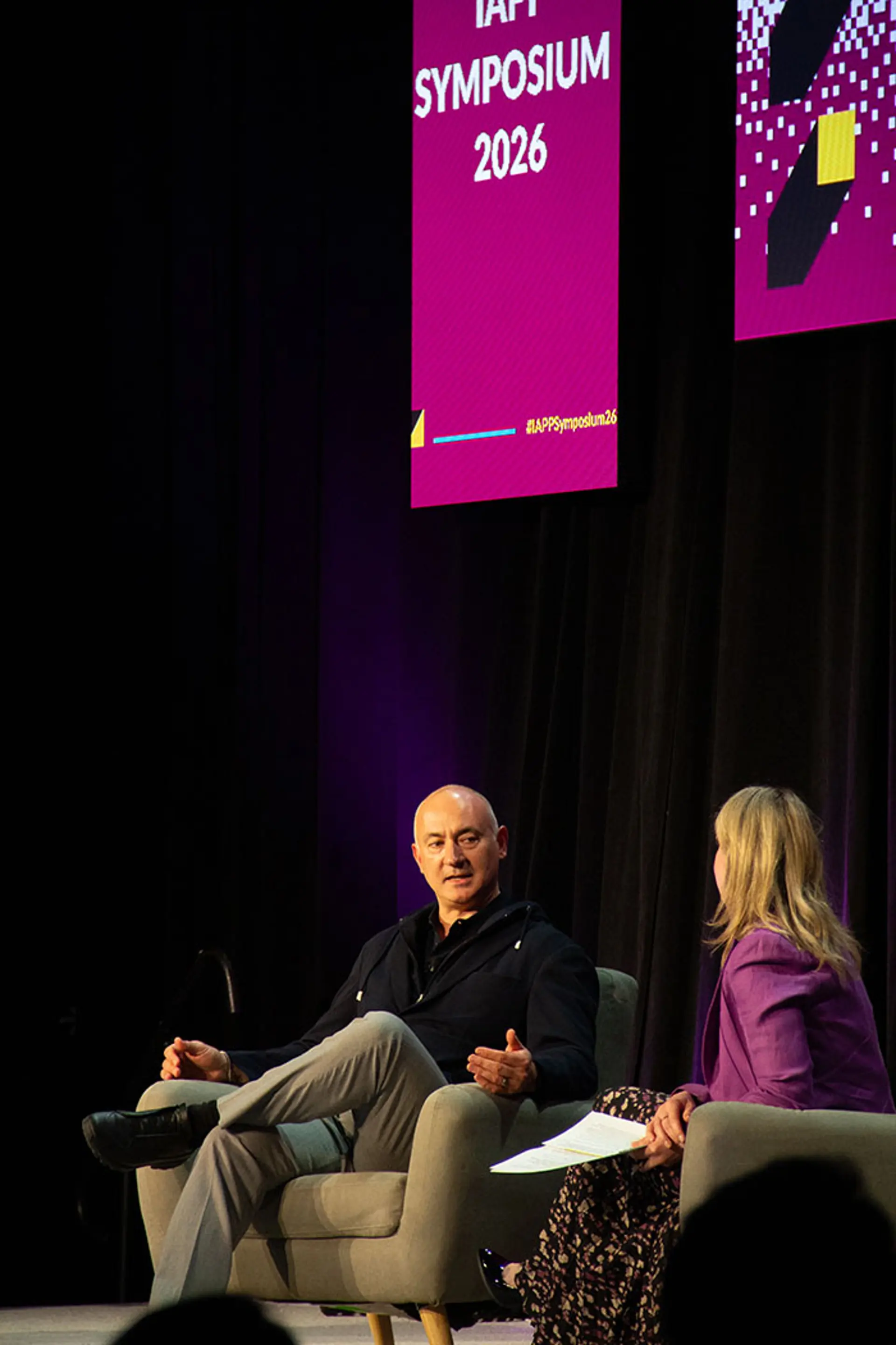 Onstage interview at IAPP Symposium 2026 featuring two speakers seated in armchairs, engaged in discussion beneath tall pink event signage.
