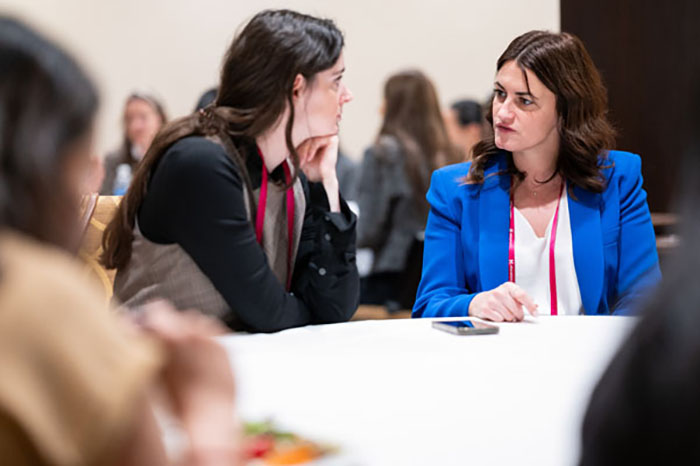 Two people seated at a round table during a networking session, engaged in conversation. A smartphone rests on the table, and other attendees are visible in the background.
