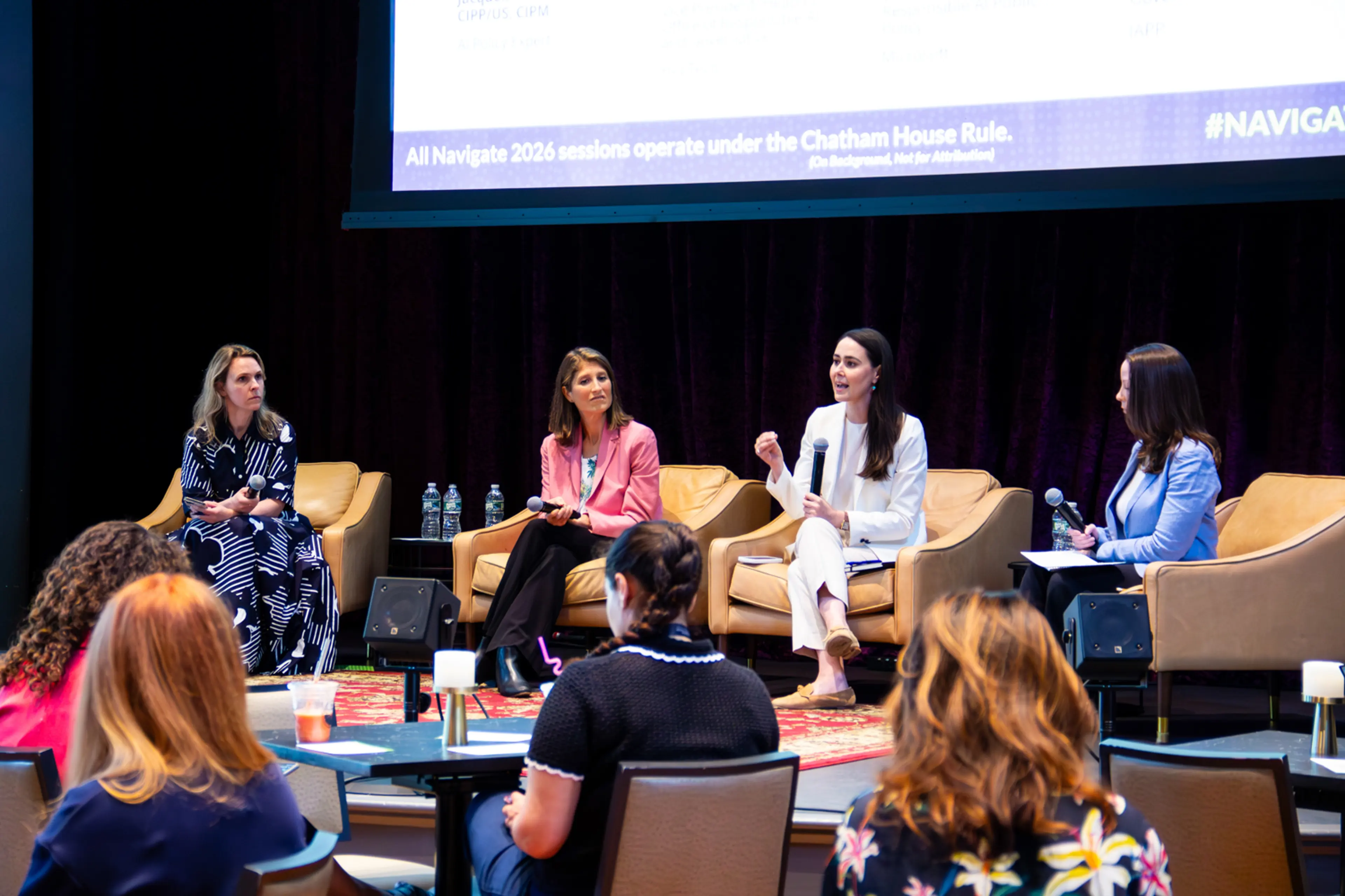 Four panelists sit on stage in armchairs during a conference discussion while audience members listen from tables in the foreground. One panelist speaks into a handheld microphone beneath a large presentation screen, and the stage is framed by dark theater curtains and soft lighting.
