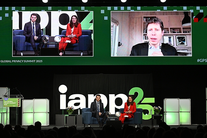 Two people seated on stage in front of a large “IAPP25” logo, with flowers on small tables. Above them, a large screen shows a split view: one side with two people seated on a couch, and the other side with a person in a home office setting. Text on the screen includes “GLOBAL PRIVACY SUMMIT 2025.”