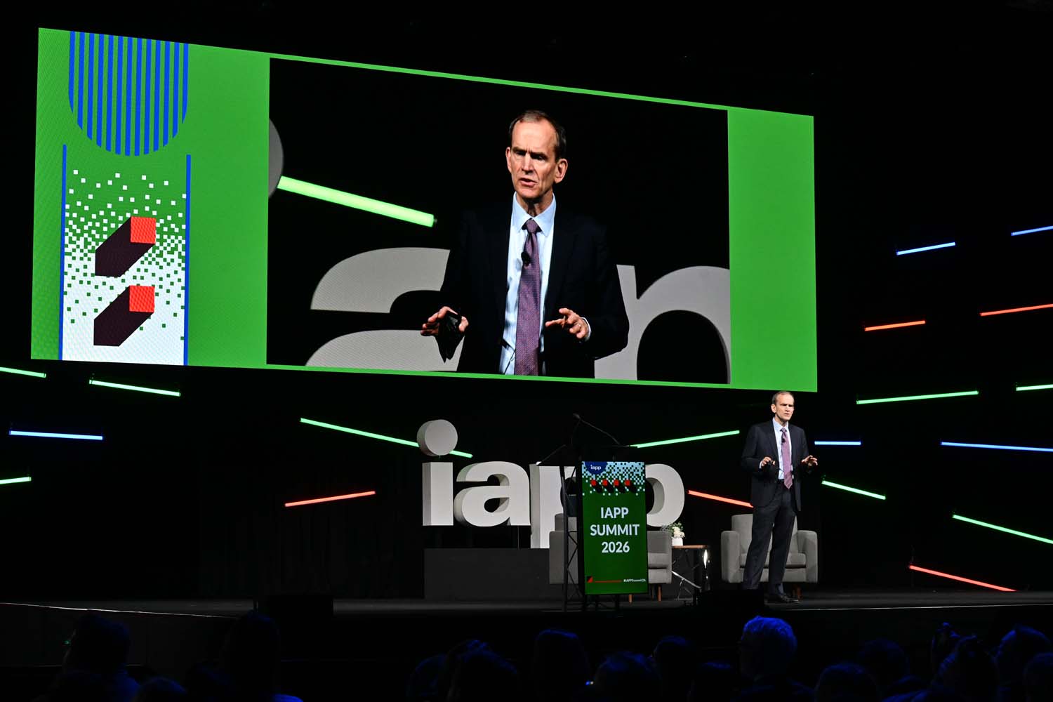 A keynote speaker stands on a large stage with a projection screen behind displaying a live close-up of the speaker, surrounded by IAPP Summit 2026 branding and colored light elements.