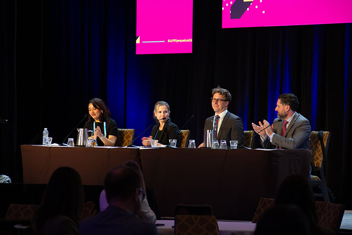 Panel of speakers seated on a stage at IAPP Symposium 2026, with microphones and water glasses on a long table as one panelist speaks and another applauds under pink event signage.