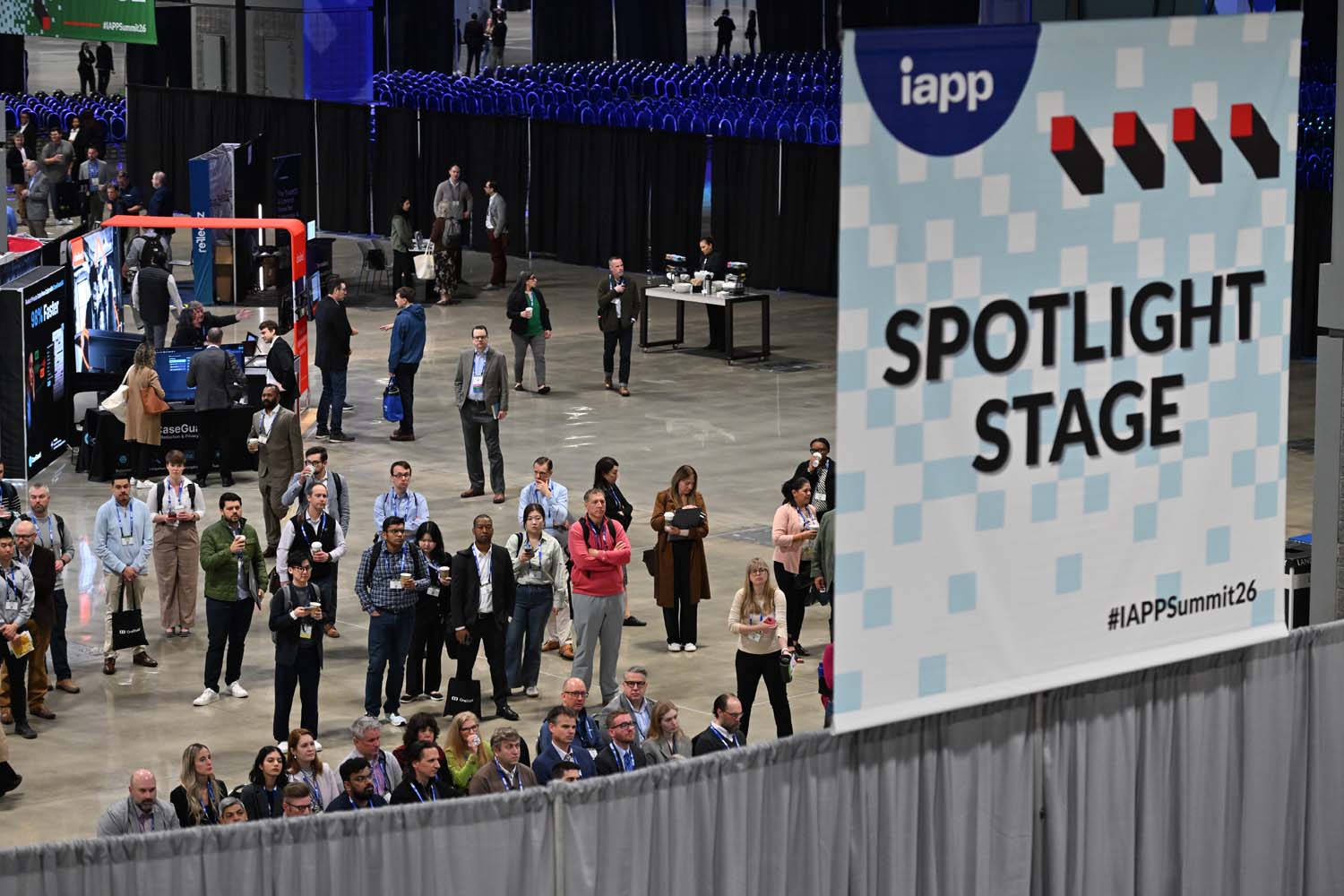 A crowd of attendees gathers on the convention floor near a tall sign reading Spotlight Stage, with display booths and standing audience members filling the open space.