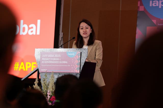 Speaker standing at a podium with signage reading “IAPP Asia 2025: Privacy Forum + AI Governance Global.” A large red screen beside the podium displays event branding and hashtag #APF25. Floral arrangements are visible at the base of the podium.