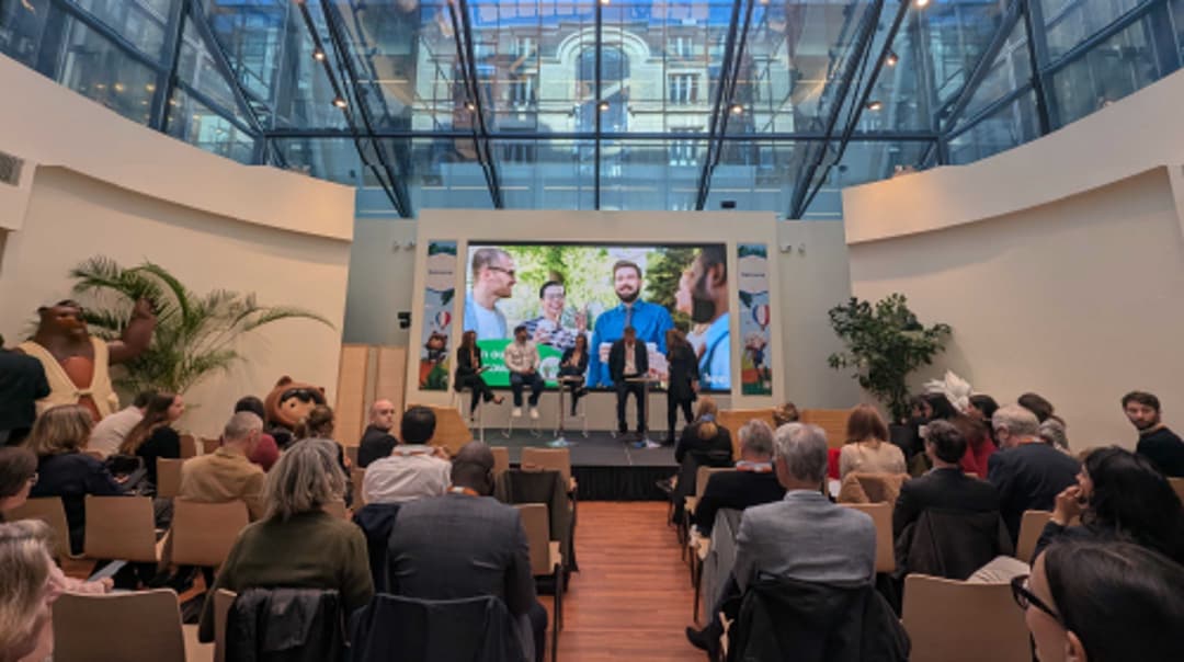 An audience seated in a glass‑roofed atrium watching a speaker and panelists onstage in front of a large screen showing an outdoor group photo.