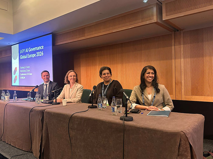 A panel of four speakers sits behind a long table on a stage with microphones, water bottles, and notes in front of them. Behind the panel, a large screen displays the title IAPP AI Governance Global Europe 2026 with event details including dates and location in Dublin.