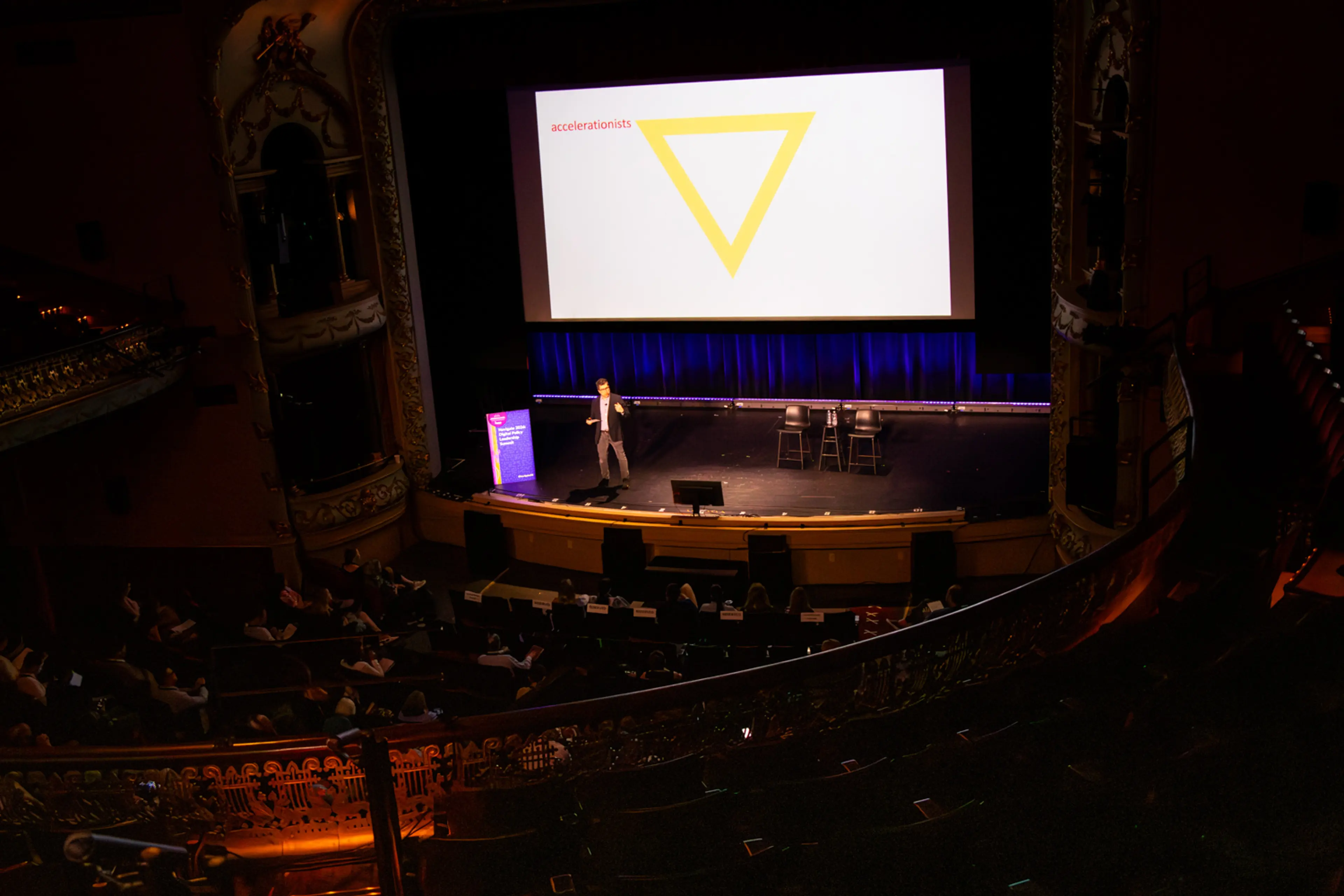 Viewed from an upper balcony, a theater audience watches a keynote presentation as a speaker stands alone on stage beneath a large screen displaying a simple slide featuring an inverted yellow triangle labeled “accelerationists.”