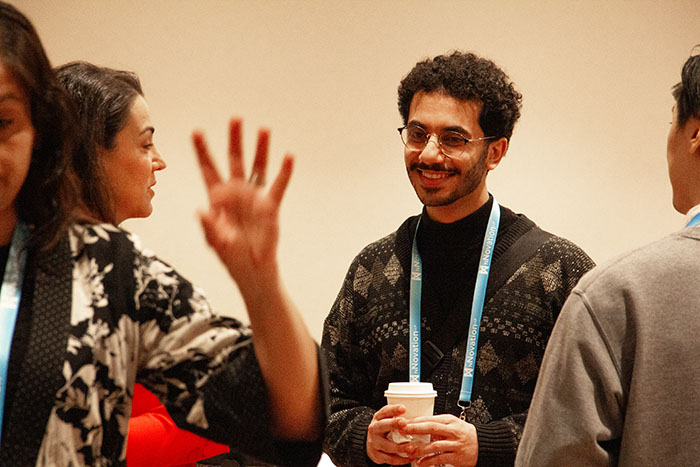 Conference attendees standing and conversing in a networking setting, with one person holding a coffee cup and wearing an event lanyard while others gesture during discussion.