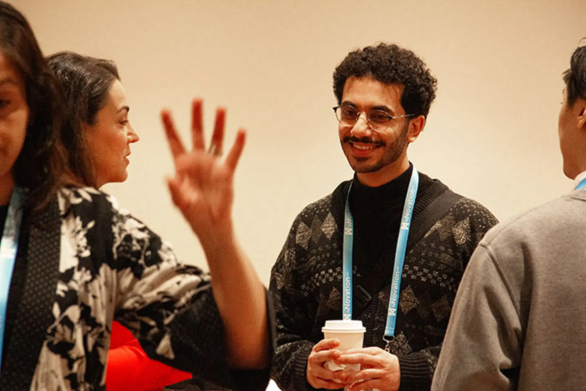 Conference attendees standing and conversing in a networking setting, with one person holding a coffee cup and wearing an event lanyard while others gesture during discussion.