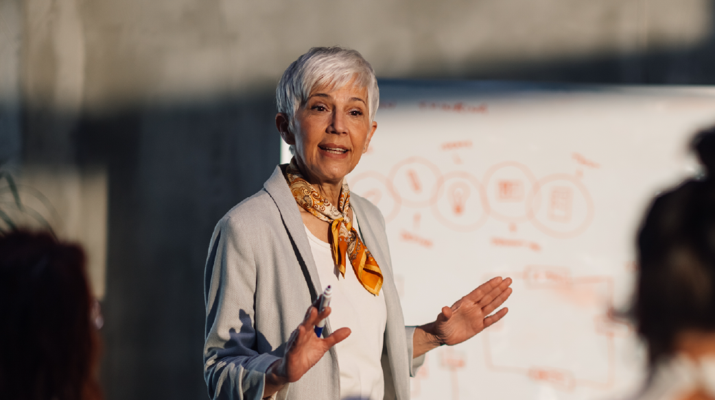 A woman in a grayish-white jacket giving a presentation.