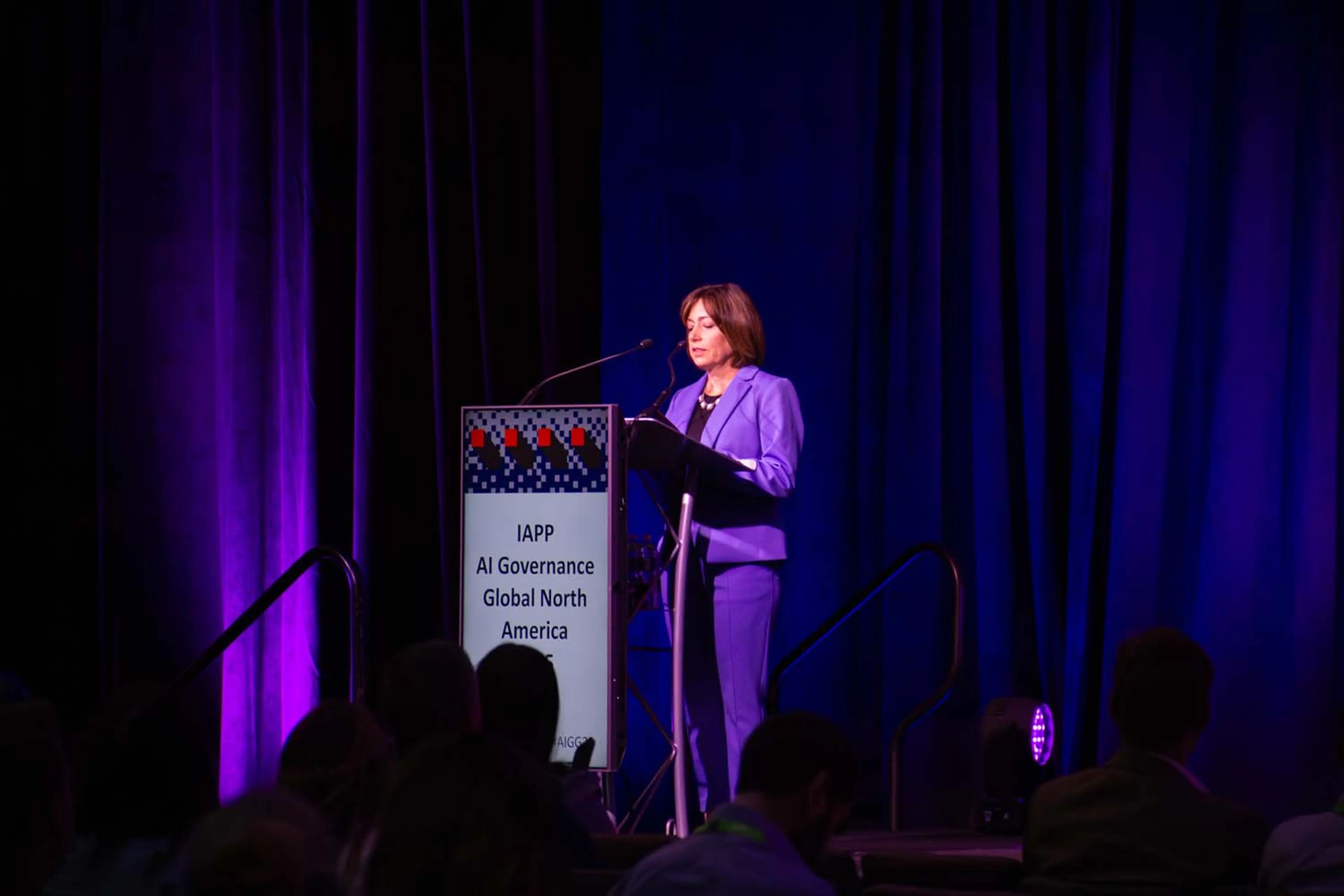 A speaker stands at a podium bearing IAPP AI Governance Global North America branding, delivering remarks on a stage framed by deep blue and purple curtains.