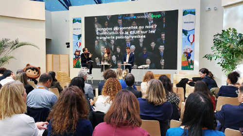 A seated audience watching panelists onstage in a bright atrium; the screen behind them displays a French‑language presentation on pseudonymized data transfers.
