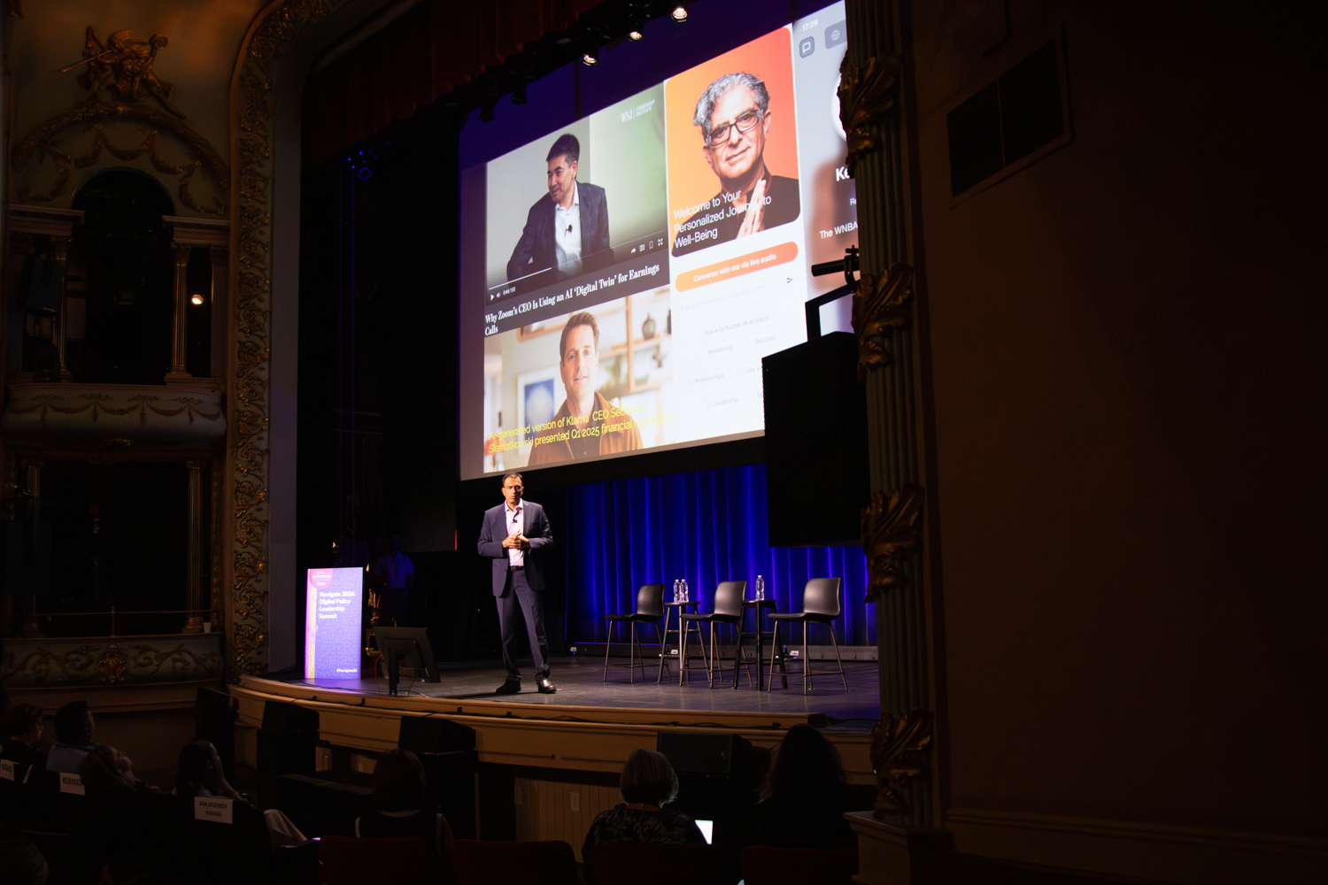 A presenter stands on the stage of an ornate theater beneath a large projection screen displaying examples of AI-generated media and digital experiences, while attendees watch from the audience and a row of empty chairs awaits a forthcoming discussion.