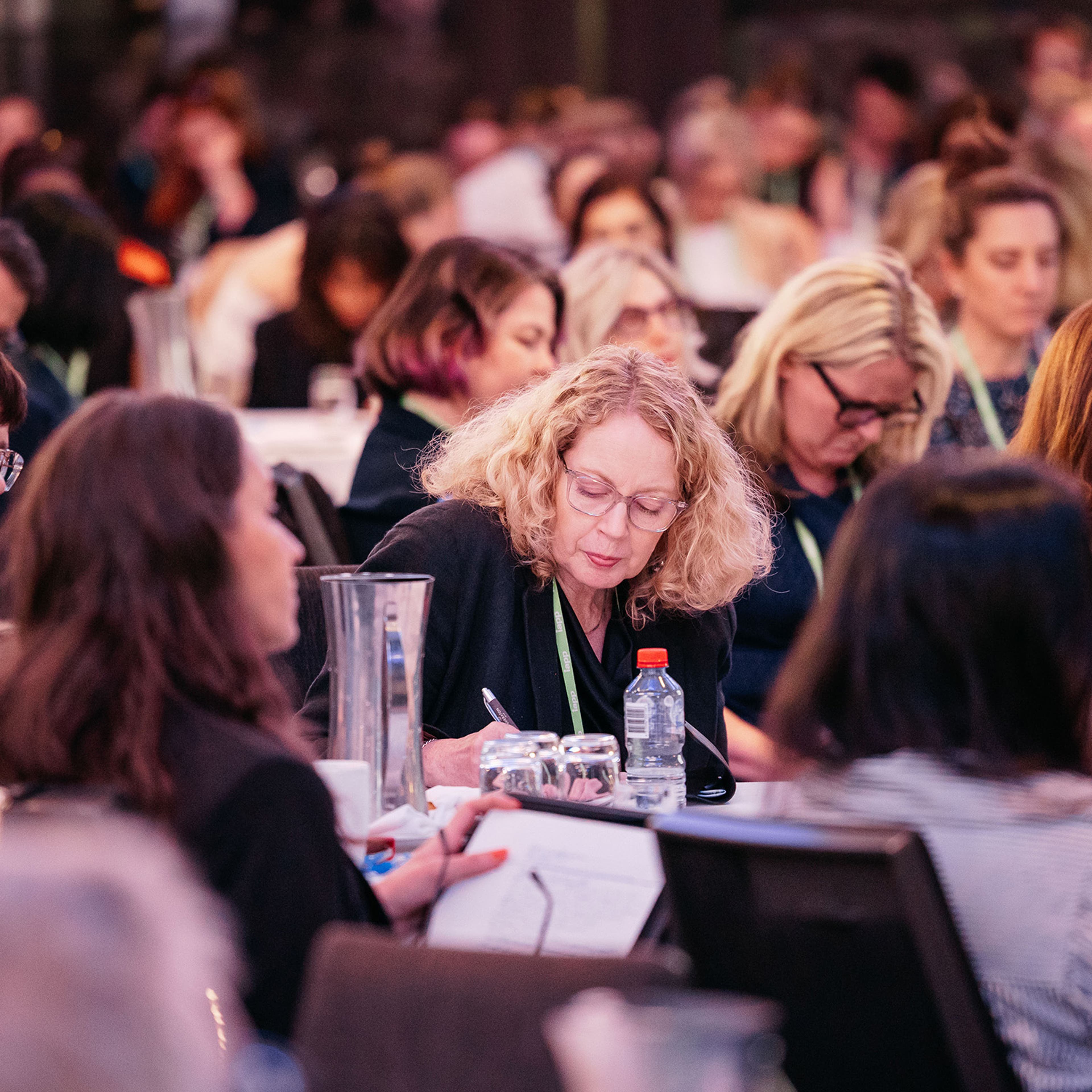 Audience seated at round tables during a conference session, taking notes and listening to speakers.