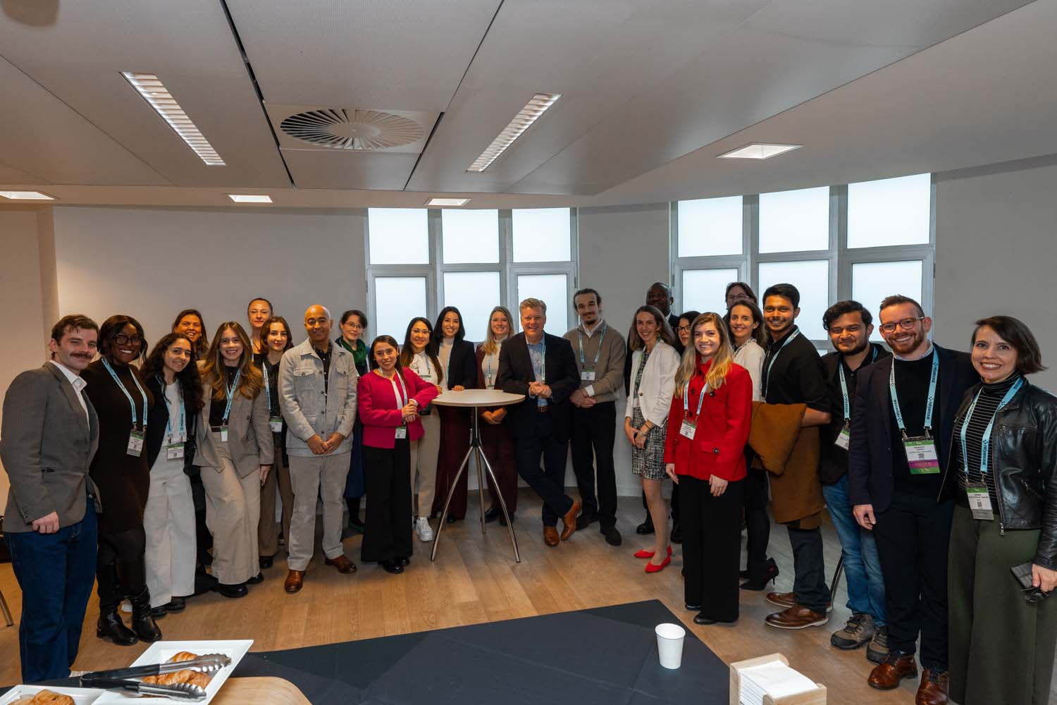 Large group of delegates posing together in a conference room with white walls and windows, standing around a tall round table.