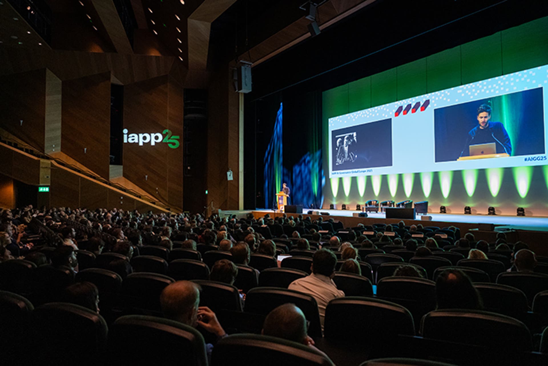 Wide view of a keynote session in a large auditorium. A speaker presents at a podium with a large screen displaying slides. Audience seated in rows.