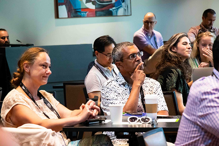 Audience seated at tables during a conference session, with notebooks, coffee cups, and lanyards visible. A colorful abstract painting hangs on the wall in the background.