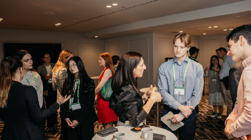People networking in a conference room, standing in small groups and talking with name badges and coffee cups visible.