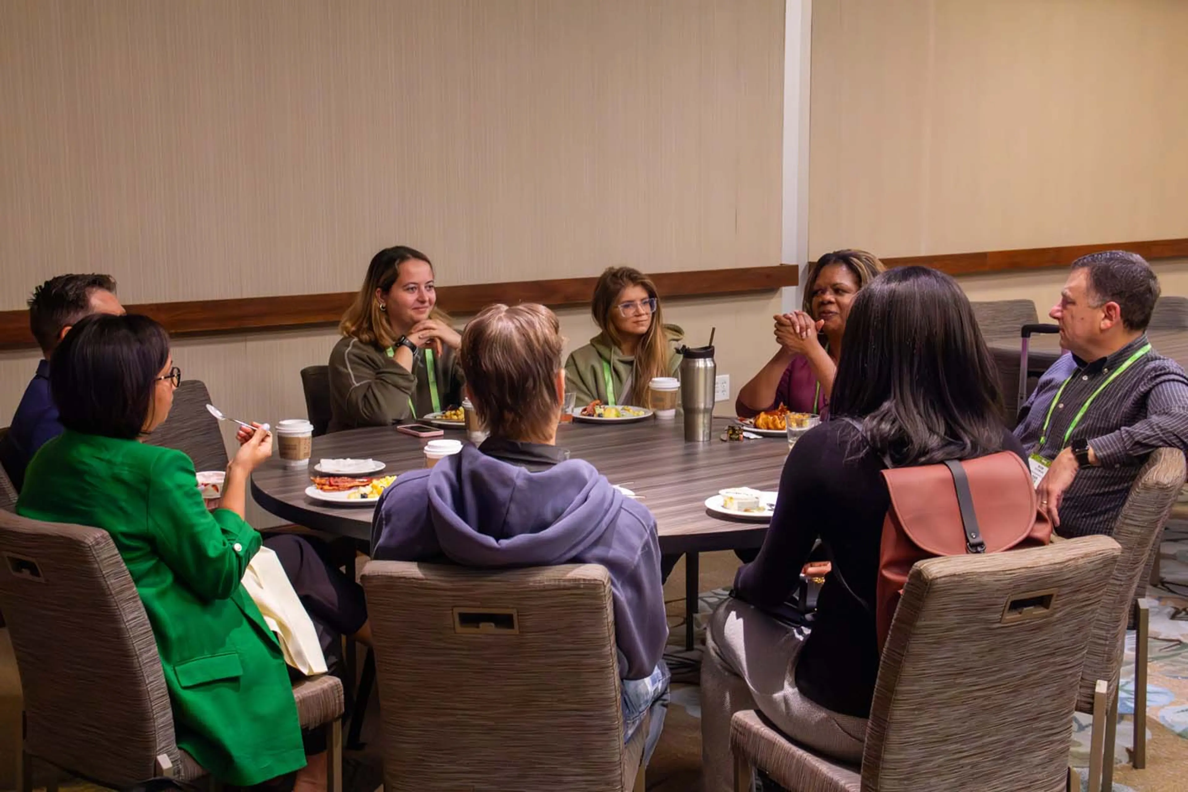 A small group of conference attendees sits closely around a round table during a meal, leaning in and conversing as plates, coffee cups, notebooks, and lanyards rest on the tabletop in a relaxed networking moment.
