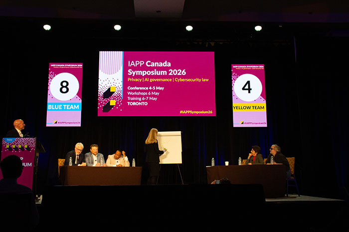 Stage session at IAPP Canada Symposium 2026 with participants seated at tables, a presenter writing on a flip chart, and large screens displaying event branding and team numbers labeled “Blue Team” and “Yellow Team”.