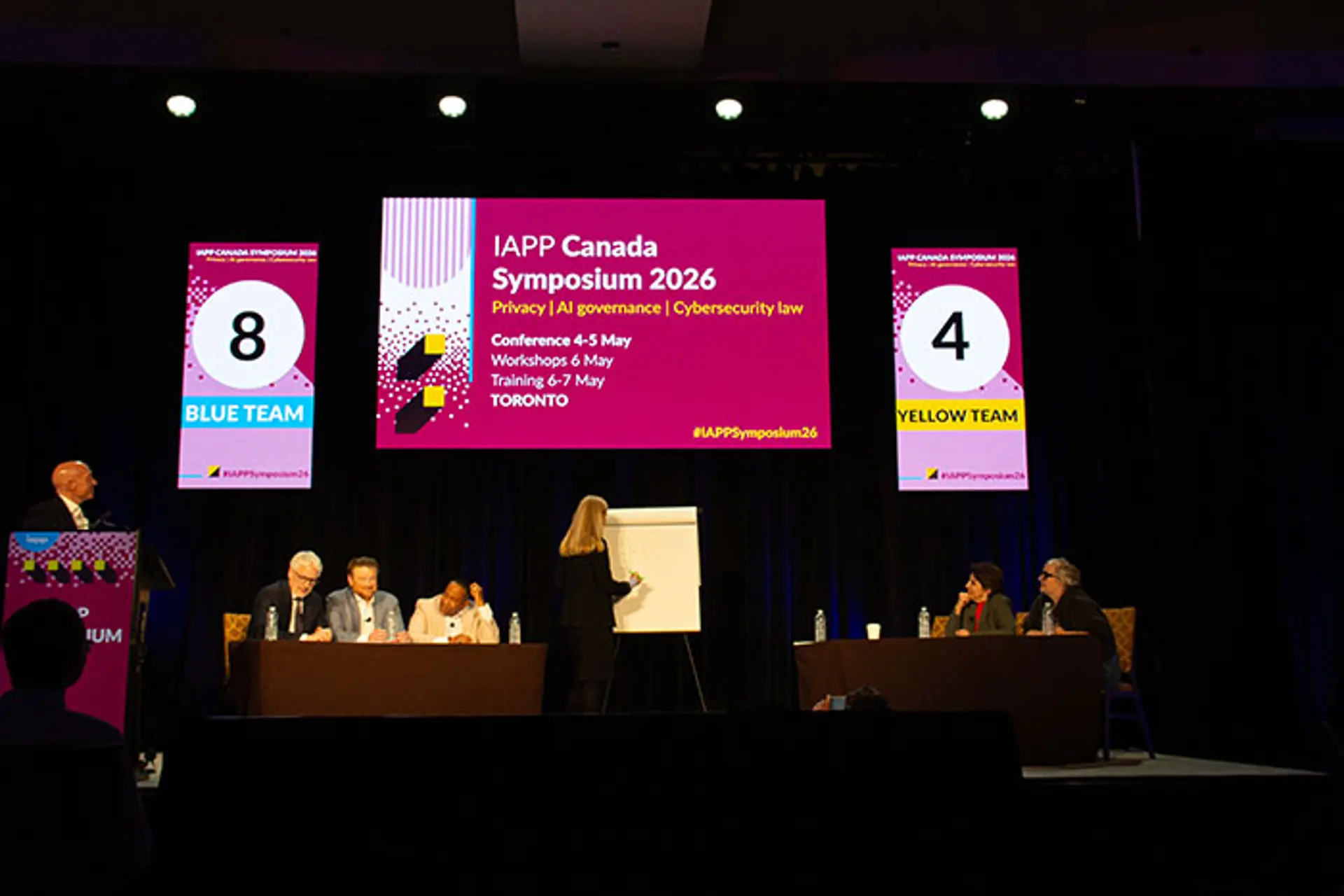 Stage session at IAPP Canada Symposium 2026 with participants seated at tables, a presenter writing on a flip chart, and large screens displaying event branding and team numbers labeled “Blue Team” and “Yellow Team”.