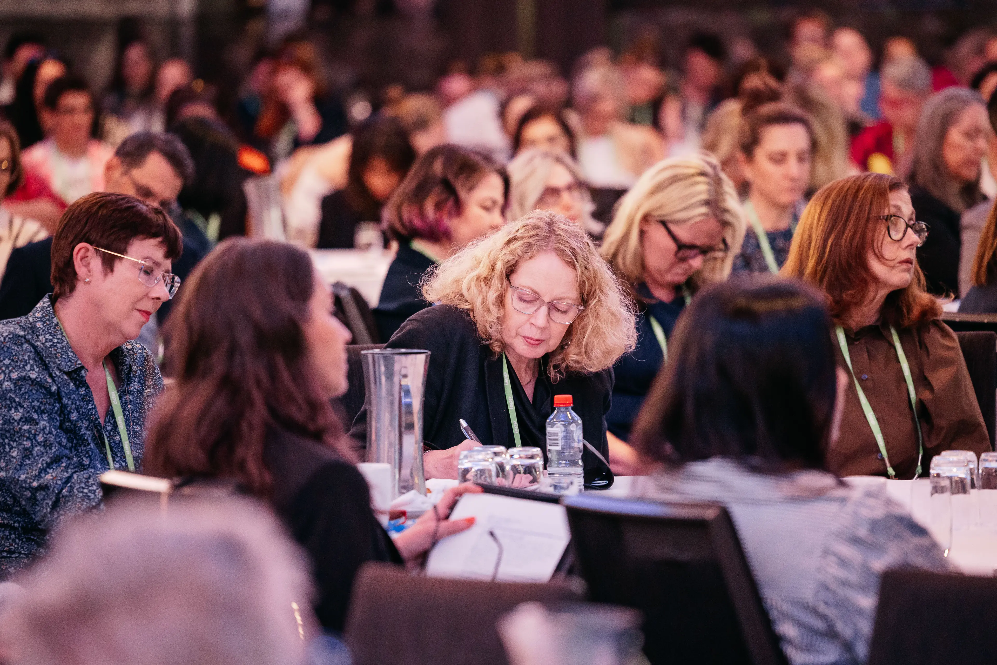 Attendees sit closely together at round tables in a large conference room, with many people wearing lanyards and writing notes while water glasses, pitchers, and notebooks are spread across the tabletops, creating a focused, studious atmosphere during a session.