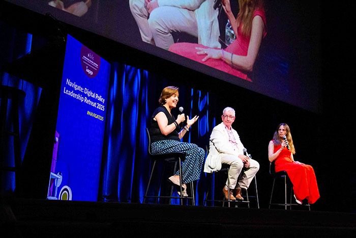 Three speakers seated on stage with microphones during a conference session. A large screen above shows the event branding: “Navigate: Digital Policy Leadership Retreat 2025.”