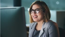 smiling woman wearing glasses and short light hair looks at a computer monitor