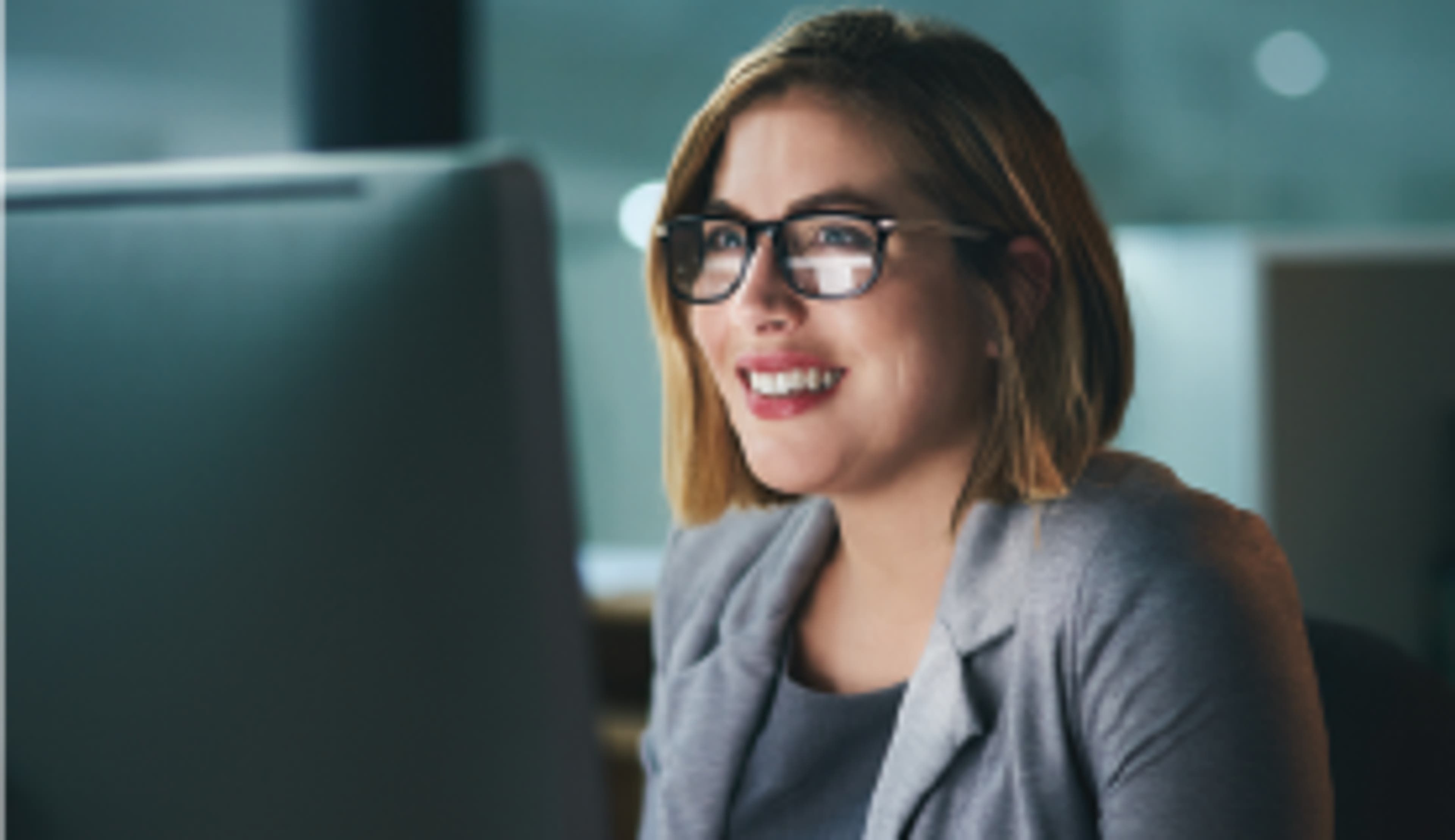 smiling woman wearing glasses and short light hair looks at a computer monitor
