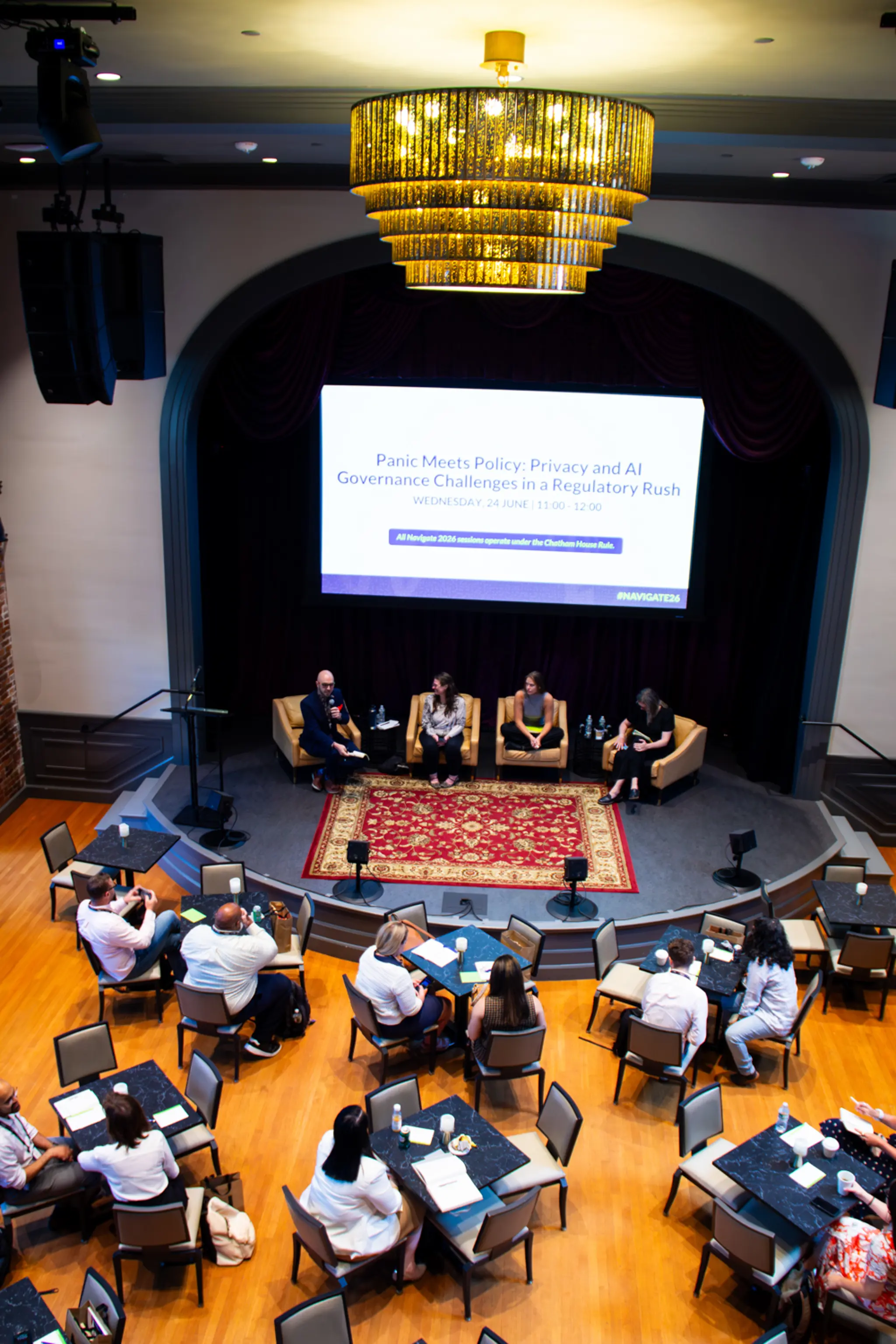 Viewed from above, attendees sit at round tables and face a stage where four speakers participate in a panel discussion beneath a large screen displaying the session title “Panic Meets Policy: Privacy and AI Governance Challenges in a Regulatory Rush,” framed by a grand chandelier and theater-style architecture.