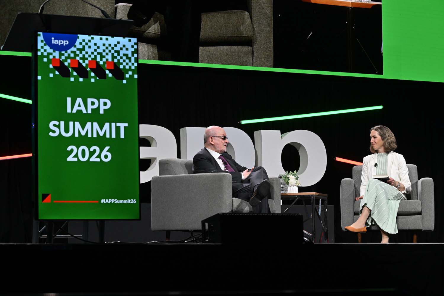 Two speakers sit facing each other on stage during an IAPP Summit 2026 session, with a small table between them and a large stage sign visible to the side.