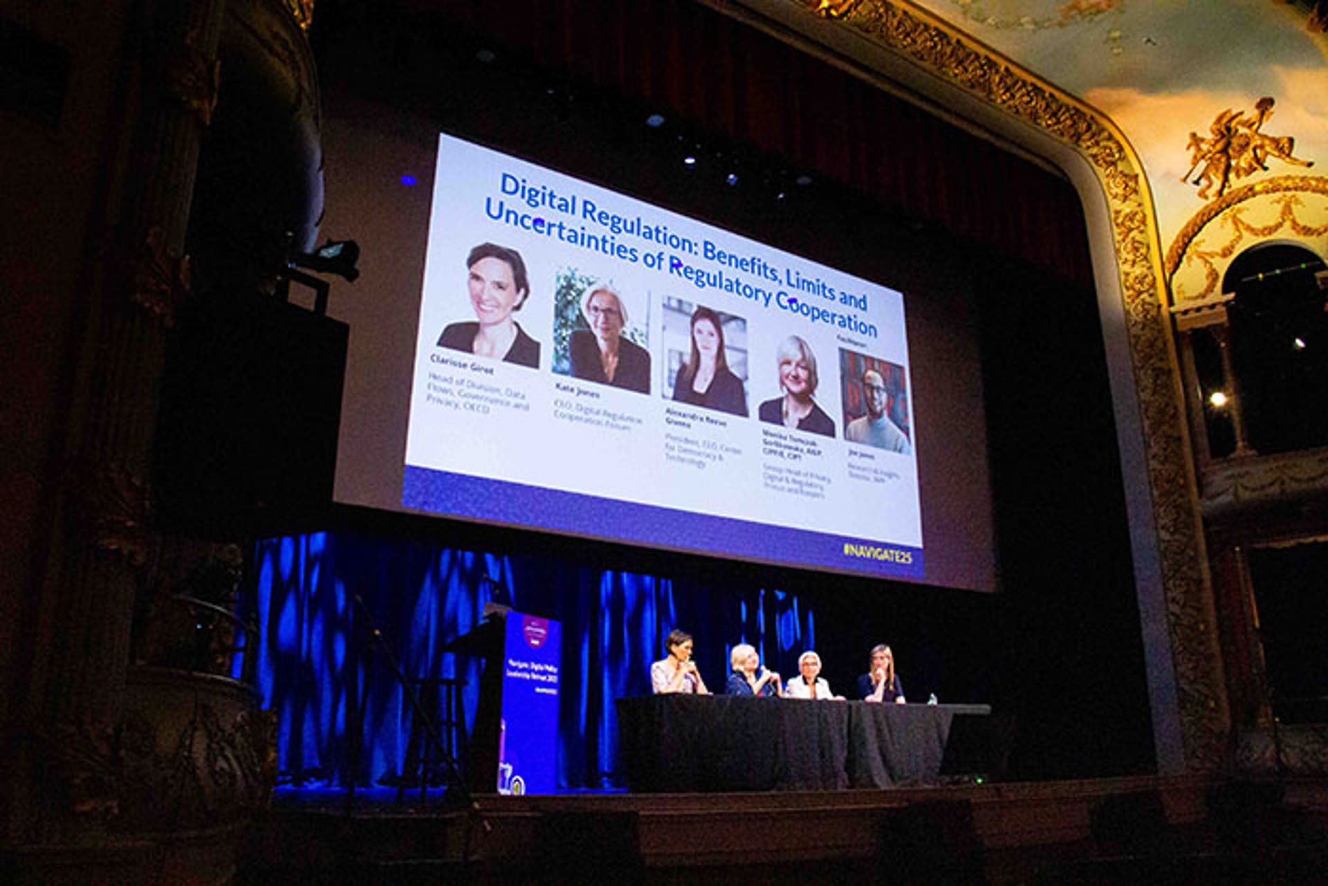 Panel discussion on a theater stage with ornate gold detailing. A large screen behind the speakers displays the session title: “Digital Regulation: Benefits, Limits and Uncertainties of Regulatory Cooperation.”