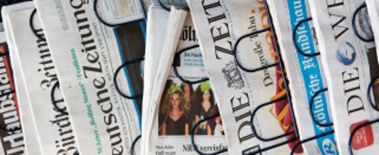 A closeup of newspapers fanned out in a stand displaying front page European headlines, primarily German newspapers.