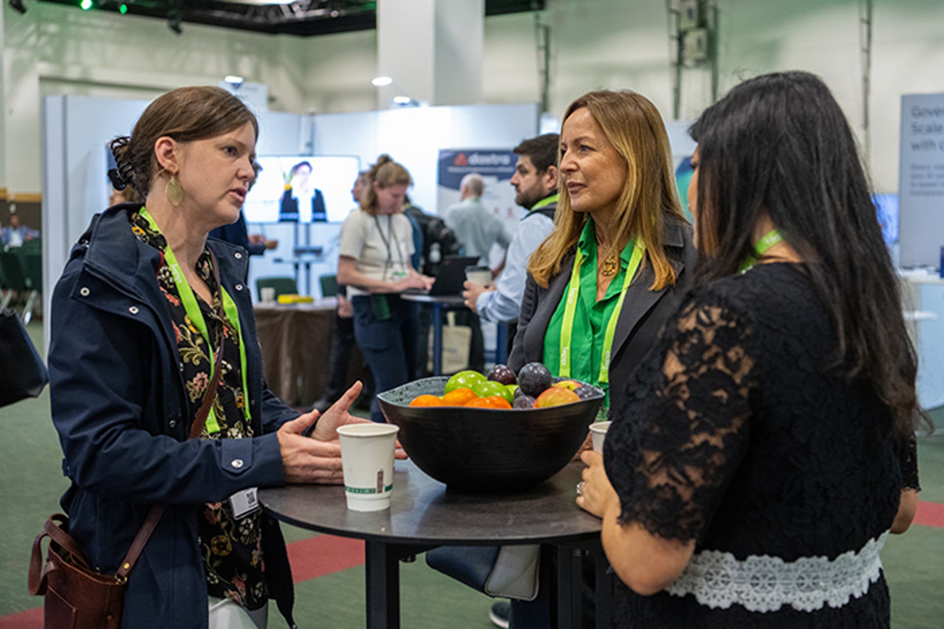Three attendees standing around a tall round table with a black bowl of colorful fruit in an exhibit hall. Booths and banners are visible in the background.