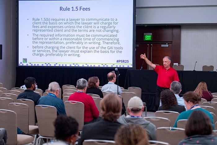 Speaker in a red shirt presenting in front of a large screen displaying text titled “Rule 1.5 Fees.” Audience members are seated in rows facing the stage.