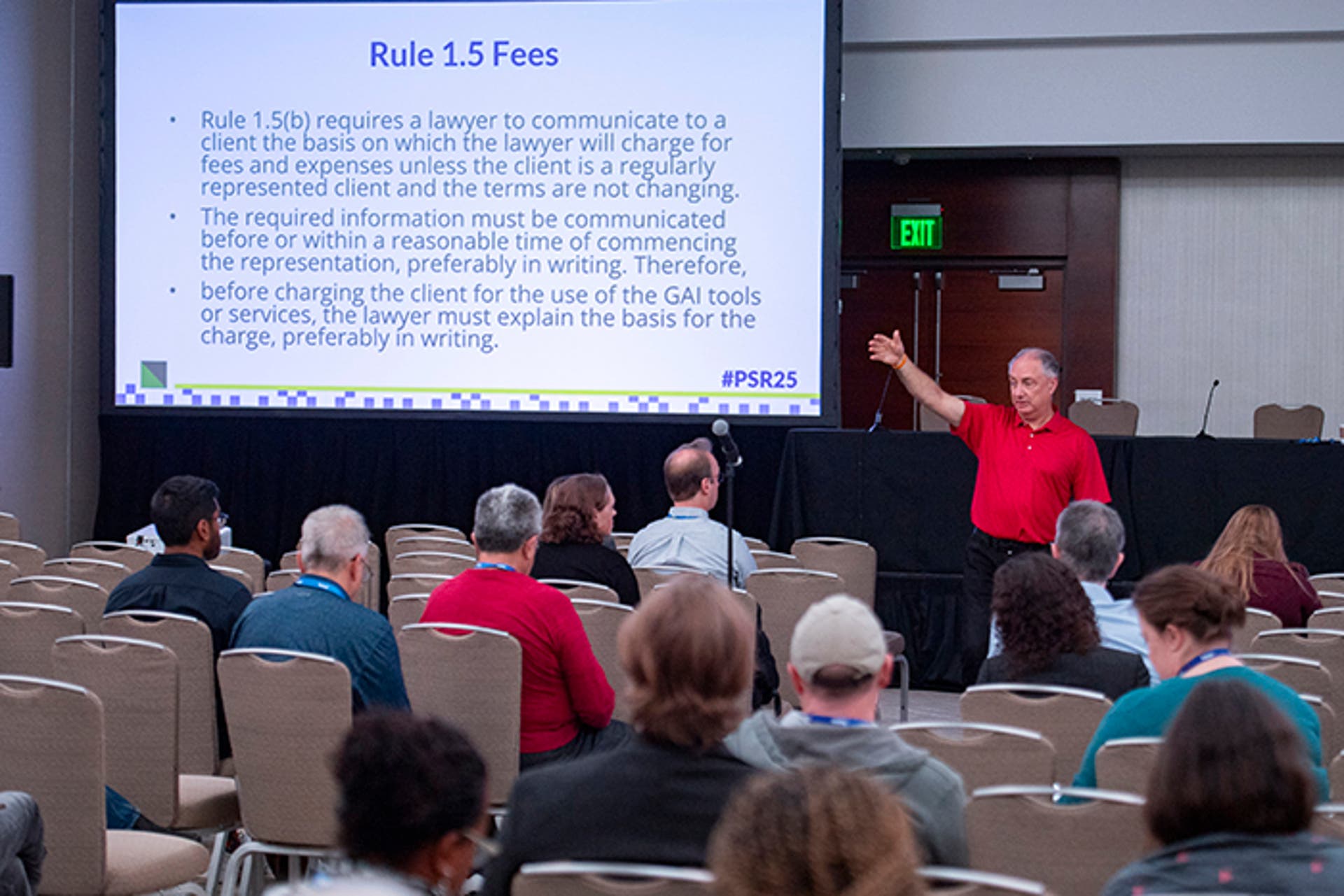 Speaker in a red shirt presenting in front of a large screen displaying text titled “Rule 1.5 Fees.” Audience members are seated in rows facing the stage.