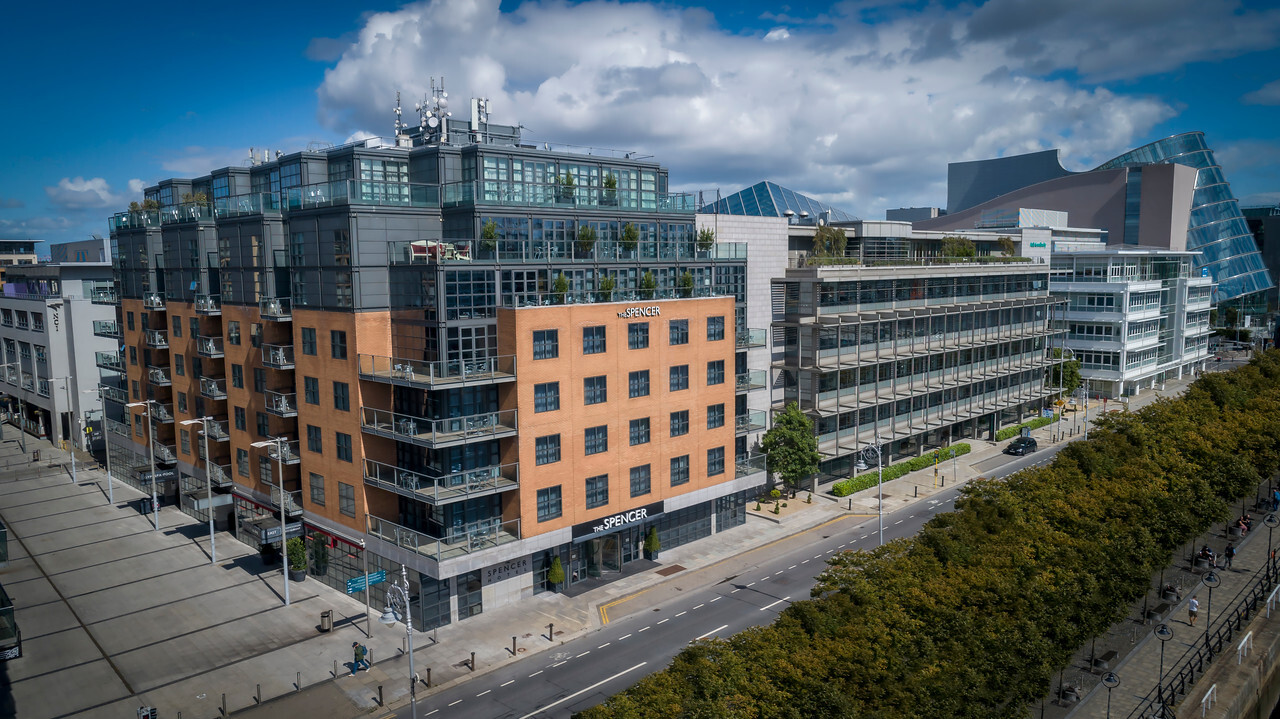 Aerial view of a modern city block with multi‑story brick and glass buildings, including the Spencer Hotel, along a tree‑lined road under a bright sky with scattered clouds.