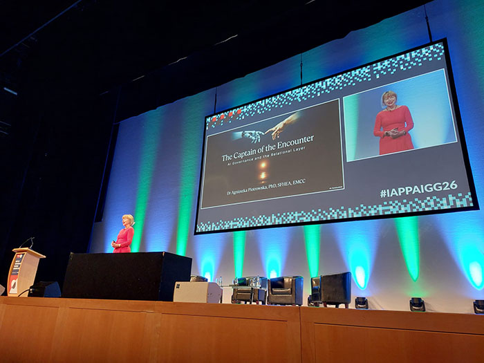 A speaker stands on a large conference stage beside a podium while a wide screen displays a presentation slide titled The Captain of the Encounter with a subtitle about governance at the international layer. Blue and green stage lighting frames the screen, and several empty chairs are arranged on the stage behind the speaker.